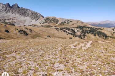 A panoramic view of a mountain landscape featuring rugged peaks and rolling hills. The foreground consists of rocky terrain with patches of grass and small shrubs, while the background showcases higher elevations with a clear blue sky and distant mountain ranges. The scene captures the natural beauty of a remote wilderness area. American Lakes Trail mountain bike trail.