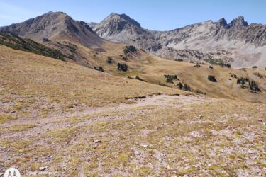 A panoramic view of a mountainous landscape featuring rocky peaks and rolling hills, with grassy and rocky terrain in the foreground. The sky is clear and blue, creating a bright contrast against the rugged mountains in the background. American Lakes Trail mountain bike trail.