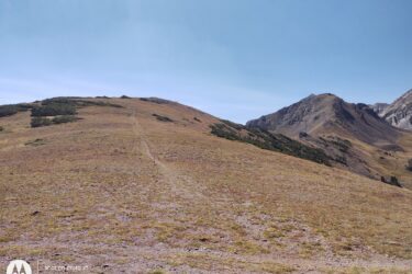 A wide view of a grassy hillside leading up to rocky mountains under a clear blue sky, with a faint trail visible winding through the terrain. American Lakes Trail mountain bike trail.