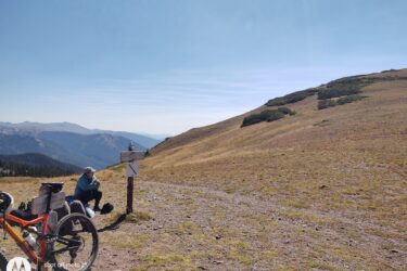 A mountainous landscape with a clear blue sky, featuring a cyclist sitting near a trail sign. In the foreground, an orange mountain bike rests on the ground, while the cyclist, wearing a light blue jacket and cap, checks their gear. The background showcases rolling hills and distant peaks, with patches of grass and sparse vegetation. American Lakes Trail mountain bike trail.