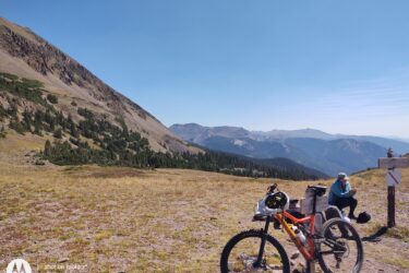 A mountainous landscape with a cyclist's orange mountain bike in the foreground. A person wearing a blue jacket sits nearby, looking at their phone. In the background, steep hills and dense evergreen trees can be seen under a clear blue sky. A trail sign is visible nearby, indicating outdoor activity in the area. American Lakes Trail mountain bike trail.