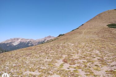 A wide view of a mountainous landscape featuring rolling hills covered in brown and green vegetation under a clear blue sky. In the background, rugged mountain peaks are visible. A narrow dirt path winds up one of the hills, suggesting a hiking trail. American Lakes Trail mountain bike trail.