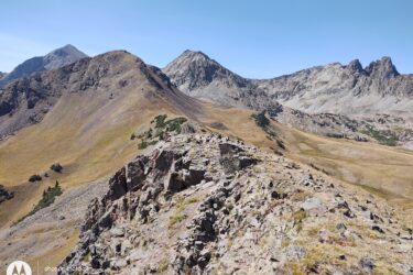 A panoramic view of rugged mountain terrain with a mix of rocky outcrops and grassland. Various peaks rise in the background under a clear blue sky, showcasing the natural beauty of the landscape. American Lakes Trail mountain bike trail.