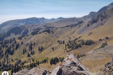 A panoramic view of a mountainous landscape featuring rolling hills covered with green and brown vegetation, dotted with conifer trees. The distant peaks are visible against a clear blue sky, and rocky formations can be seen in the foreground. American Lakes Trail mountain bike trail.