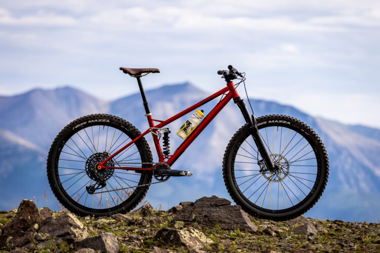 A red mountain bike positioned on rocky terrain, set against a backdrop of mountains under a cloudy sky. The bike features wide tires, a suspension system, and a water bottle attached to the frame.