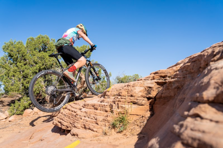 A mountain biker skillfully navigating over a rocky terrain during a sunny day, with clear blue skies and green shrubs in the background.