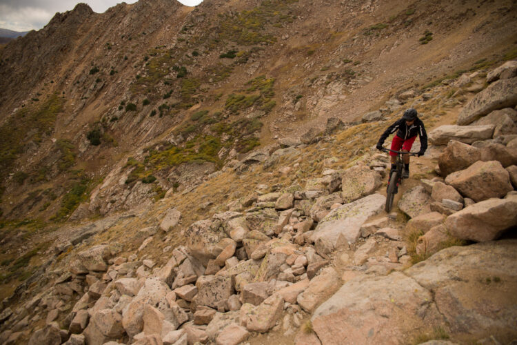 A mountain biker navigates a rocky trail on a mountainous terrain, surrounded by steep slopes and sparse vegetation. The biker is wearing a helmet and riding gear, with a focus on the challenging landscape filled with large stones and gravel.