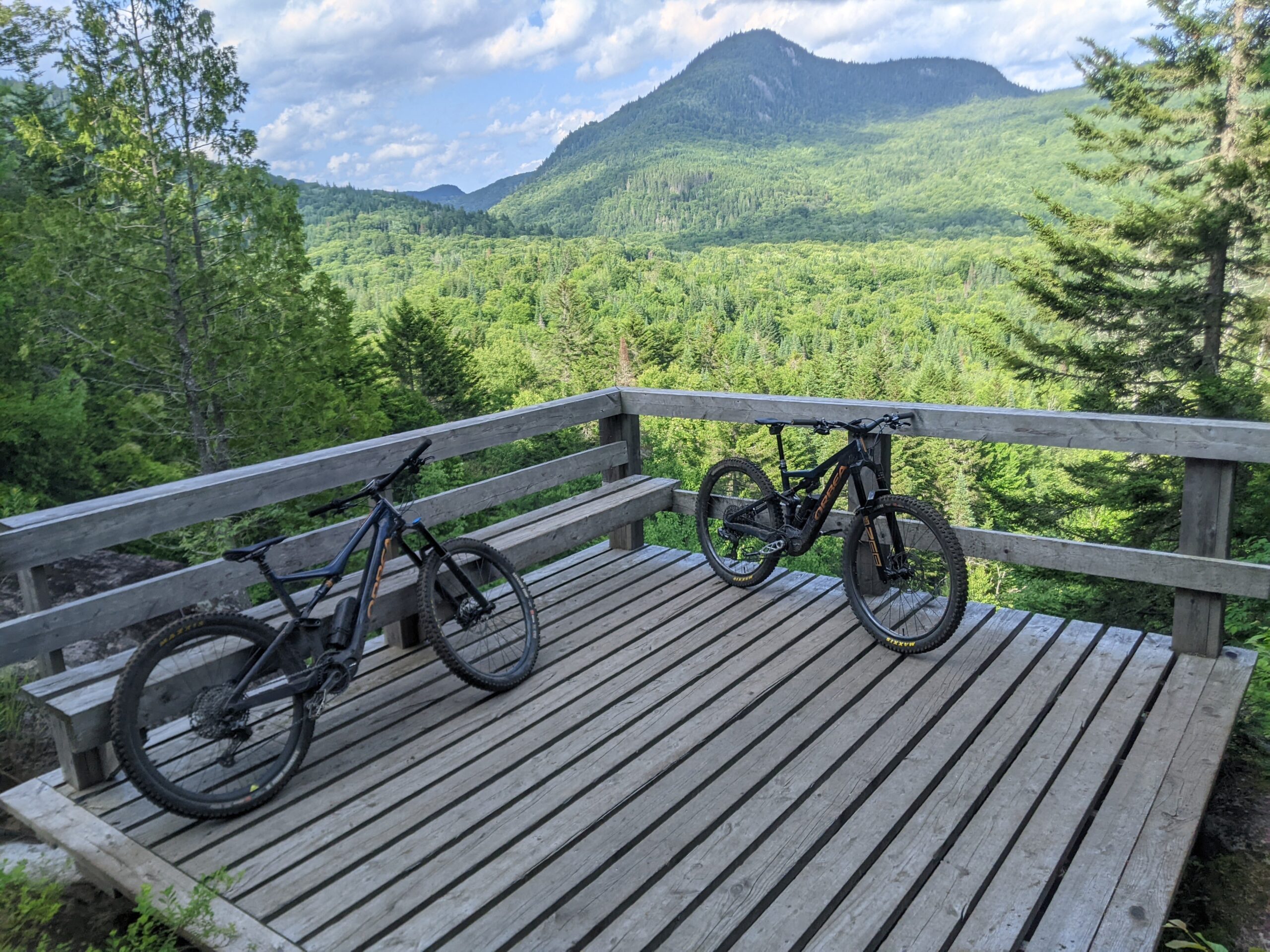 Two mountain bikes parked on a wooden viewing platform overlooking a lush green valley surrounded by hills and trees under a partly cloudy sky. Legende mountain bike trail.