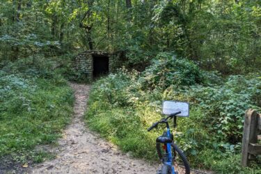 A blue bicycle parked on a sandy path leading to a stone structure partially hidden in dense green foliage in a forested area. A sign is positioned near the bike, providing information about the location. Sunlight filters through the trees, illuminating the scene. Potawatomi trail mountain bike trail.