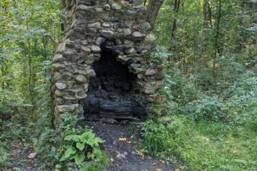 A weathered stone chimney stands amidst a lush green forest, partially overgrown with foliage. The structure features a rounded arch opening and is surrounded by grass and scattered fallen leaves, highlighting its rustic charm and the natural setting. Potawatomi trail mountain bike trail.
