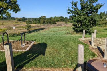 A sunny outdoor scene featuring a grassy field with scattered trees in the background. In the foreground, there are two exercise or fitness stations made of black metal beside a surface covered in wood chips. A sign can be seen in the distance, and the landscape is clear with a blue sky overhead. Sharon Mills MTB Trails mountain bike trail.
