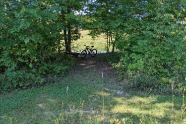 A scenic view of a pathway leading through lush green foliage, with two bicycles resting on the side. The background features a serene body of water, partially obscured by trees. Sunlight filters through the leaves, creating a peaceful outdoor atmosphere. Sharon Mills MTB Trails mountain bike trail.