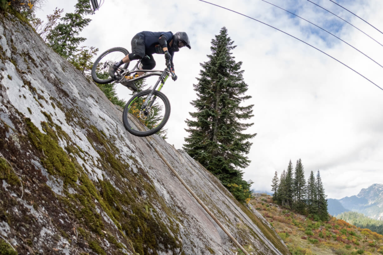 A mountain biker rides down a steep, rocky surface surrounded by pine trees, with a cloudy sky above and hills in the background. The cyclist is wearing protective gear and is captured in motion, showcasing a dynamic biking maneuver.