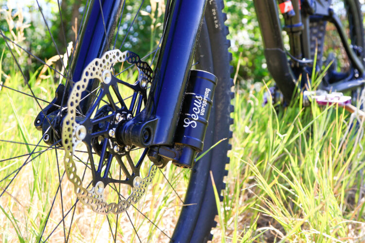 A close-up view of a mountain bike's front wheel, showcasing the disc brake and suspension component. The bike is set against a backdrop of tall grasses and greenery, illustrating an outdoor setting.