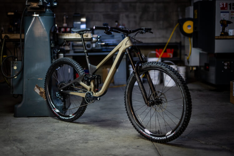 A modern mountain bike in a workshop setting, featuring a sleek frame in a light tan color and black accents. The bike has robust tires designed for off-road terrain and is positioned on a concrete floor with various tools and equipment in the background. Soft lighting highlights the bike's design and details.