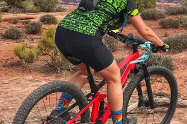 A person riding a red mountain bike on a rocky terrain, wearing a green patterned shirt and black shorts, with a helmet and a backpack. The background features a scenic view of mountains and cloudy skies. Getaway mountain bike trail.