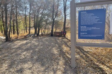 Trail entrance at Quarry Lake Park featuring a sign with trail rules and safety tips, surrounded by trees and fallen leaves. A distant view of a trailhead and nearby signage indicating emergency contact information. Xcel Energy Mountain Bike Park mountain bike trail.