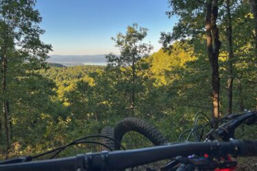A mountain bike is positioned in the foreground, partially obscuring a scenic view of a lush green forest and a distant lake, under a clear blue sky. Miller's Goat Trail mountain bike trail.