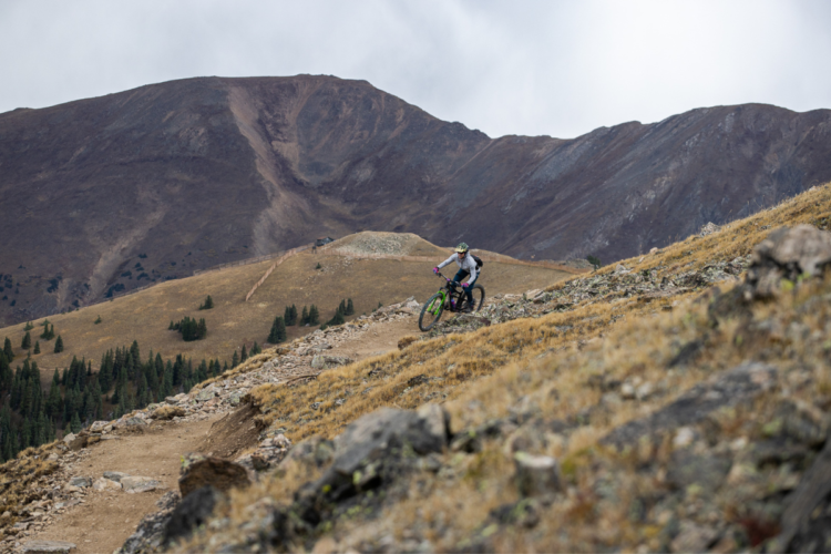 A mountain biker navigating a rocky, steep trail in a mountainous landscape with rolling hills and sparse vegetation under a cloudy sky.