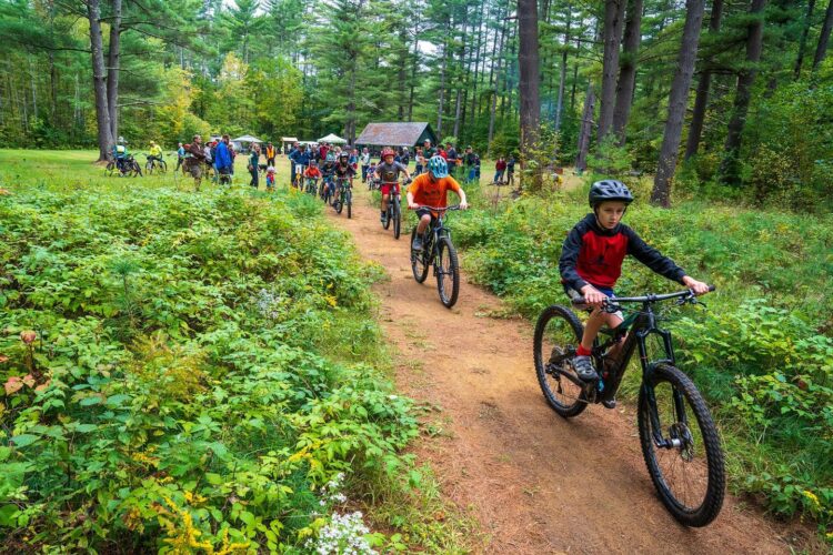 A group of children and adults riding bicycles on a dirt path through a green, wooded area. They are surrounded by tall trees and bushes, with a few people walking along the path. A cabin and tents can be seen in the background, suggesting a recreational outdoor event.