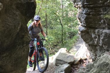 A mountain biker navigating a rocky trail between two large boulders, surrounded by lush green trees and sunlight filtering through the foliage. The cyclist is wearing a helmet and sports gear, and appears focused on the path ahead. Devil's Den State Park mountain bike trail.