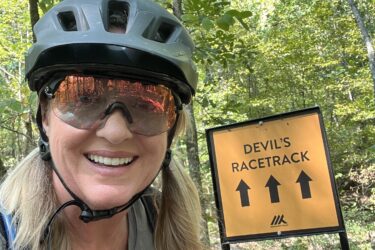 A smiling person wearing a gray helmet and sunglasses, posing in front of a trail sign that reads "Devil's Racetrack" with arrows pointing upward. The background features a lush green forest with trees and foliage. Devil's Den State Park mountain bike trail.