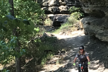 A cyclist navigating a rocky trail surrounded by lush greenery and a natural rock overhang. Devil's Den State Park mountain bike trail.