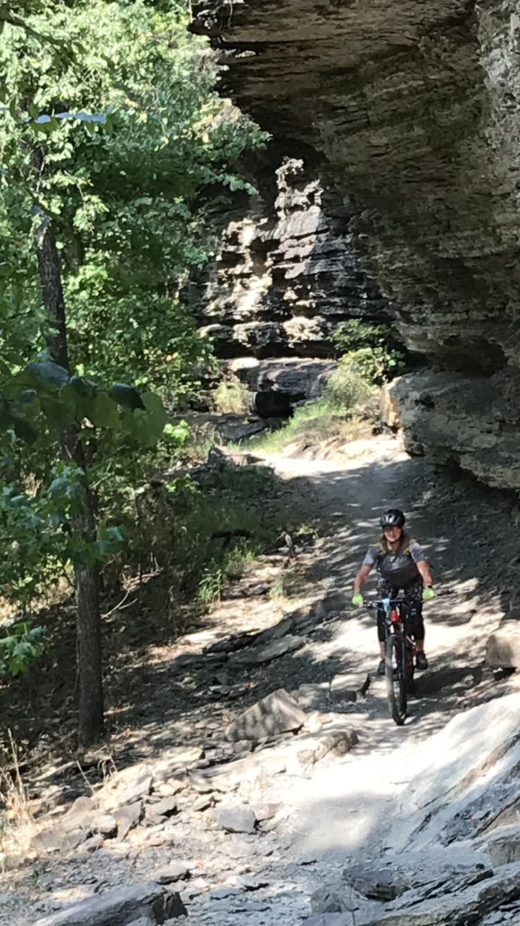 A cyclist navigating a rocky trail surrounded by lush greenery and a natural rock overhang. Devil