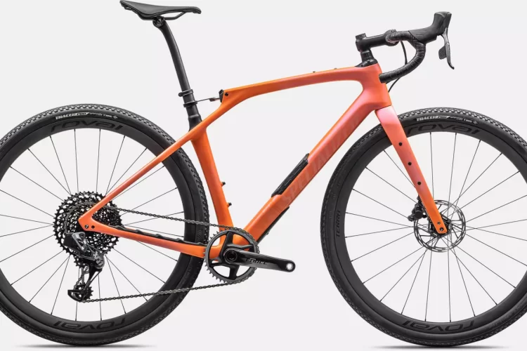 A sleek, modern bicycle featuring a gradient orange frame, disc brakes, and wide tires designed for gravel or off-road riding. The bike is positioned against a white background, showcasing its lightweight design and advanced features.