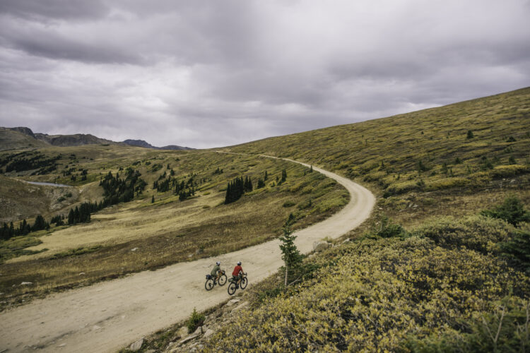 Two mountain bikers ride along a winding dirt path through a grassy hillside, with a backdrop of rolling hills and cloudy skies. The landscape is dotted with sparse trees and shrubs, showcasing an inviting but rugged outdoor setting.