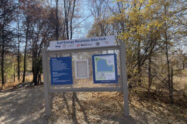Sign at the entrance of the Xcel Energy Mountain Bike Park, displaying trail rules, safety tips, and a park map. Surrounding the sign are trees with autumn foliage and a gravel path leading into the park. Xcel Energy Mountain Bike Park mountain bike trail.