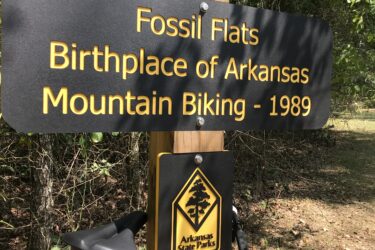 Sign marking "Fossil Flats, Birthplace of Arkansas Mountain Biking - 1989," with an Arkansas State Parks logo, positioned alongside a mountain bike. The background features natural foliage and wooded area. Devil's Den State Park mountain bike trail.