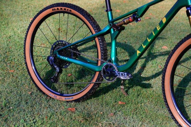 A close-up image of a mountain bike showcasing its green frame with "ALLIED" branding, featuring brown Maxxis tires, a visible drivetrain with an X01 derailleur, and a combination of gear components. The bike is positioned on grass, highlighting its robust design and outdoor setting.