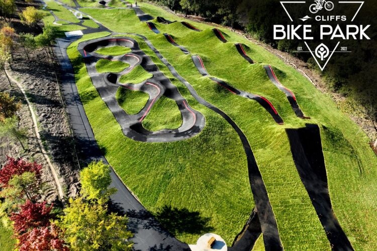 Aerial view of Cliffs Bike Park, featuring a vibrant green bike track with various hills and curves designed for biking. The paths are marked with black asphalt and red outlines, surrounded by lush grass and trees. The park logo is displayed in the corner.