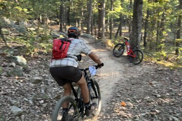 A mountain biker, wearing a helmet and a red backpack, is riding along a dirt trail in a wooded area. Sunlight filters through the trees, casting dappled shadows on the ground. A second bicycle, red in color, is parked off to the side on the trail. The terrain is rocky and surrounded by lush green foliage. Miller's Goat Trail mountain bike trail.