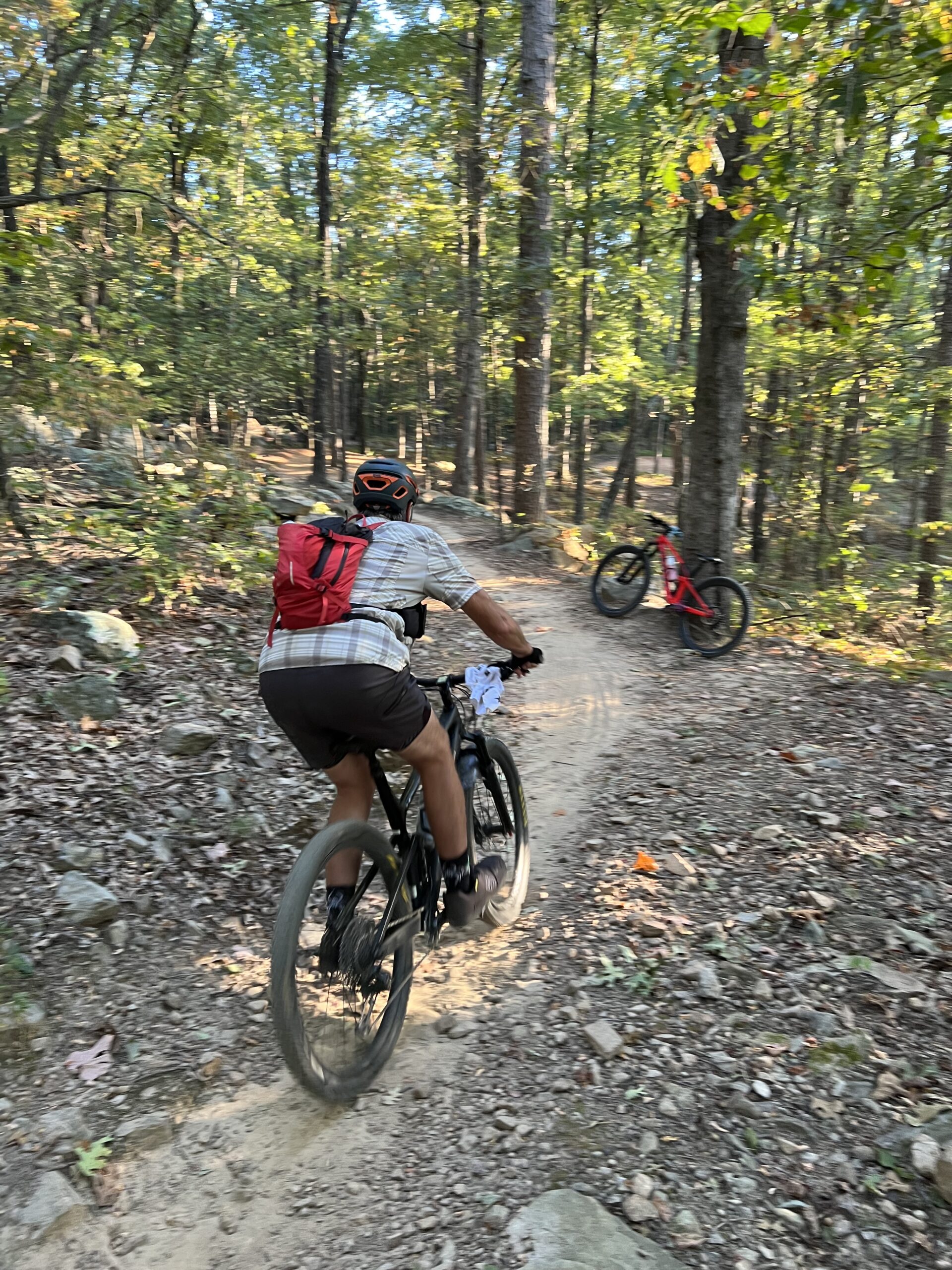 A mountain biker, wearing a helmet and a red backpack, is riding along a dirt trail in a wooded area. Sunlight filters through the trees, casting dappled shadows on the ground. A second bicycle, red in color, is parked off to the side on the trail. The terrain is rocky and surrounded by lush green foliage. Miller