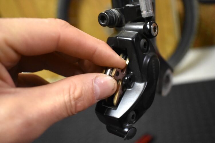 A close-up image of a person's hand adjusting or repairing a bike component, specifically focusing on the attachment mechanism of a bicycle derailleur. The hand is gently manipulating a metal piece against a black metal body, with tools and a blurred background suggesting a workshop setting.
