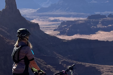 A mountain biker stands on the edge of a rocky canyon, gazing out over a vast landscape of red rock formations and deep valleys under a clear blue sky. The bike is parked beside them, and the scene captures a sense of adventure and appreciation for nature. Dead Horse Point State Park mountain bike trail.