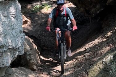 A cyclist navigating a rocky trail beneath a rocky overhang in a wooded area, surrounded by natural terrain and dappled sunlight. Devil's Den State Park mountain bike trail.