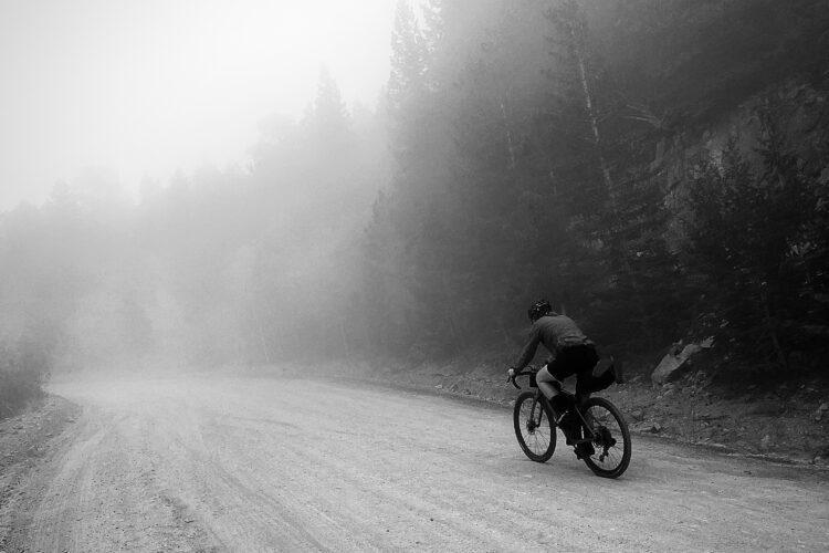 A cyclist riding on a dirt road through a foggy forest, surrounded by trees in a monochrome setting. The cyclist is leaning forward, navigating a curve in the road as the mist creates a mysterious atmosphere.