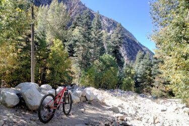 A mountain bike resting on a trail surrounded by lush trees and rocky terrain, with steep mountain slopes in the background under a clear blue sky. Little Cottonwood Pipeline mountain bike trail.