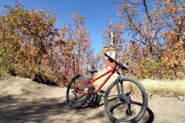 A mountain bike rests on a dirt trail with vibrant autumn foliage in the background. A wooden sign marked "Hollow" and additional warning signs are visible nearby, indicating trail guidelines. The sky is clear and blue, highlighting a sunny day. Canyon Hollow mountain bike trail.