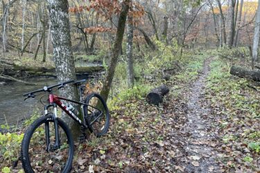 A mountain bike leaning against a tree beside a winding dirt trail, surrounded by autumn foliage and a small stream. The path is covered in fallen leaves, leading into a serene forested area. Fort Defiance mountain bike trail.