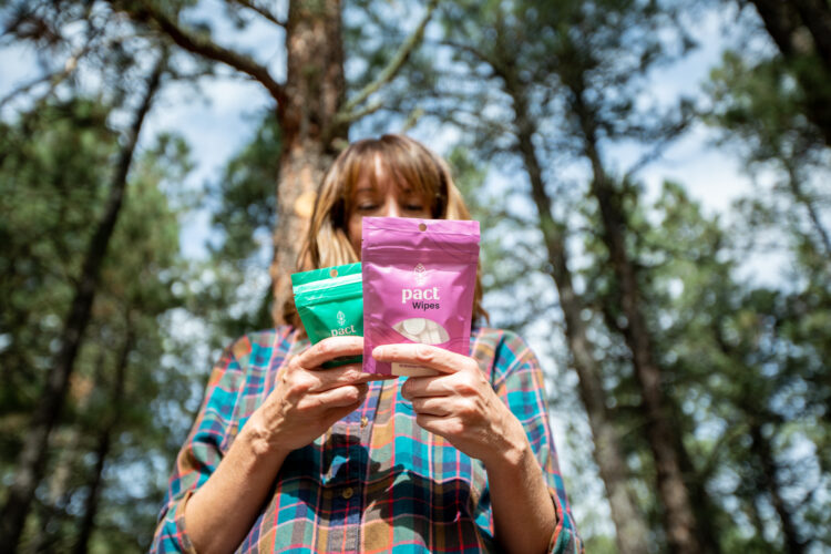 A person outdoors in a forest holds two packets of pact wipes—one green and one pink—while examining their contents. The individual is wearing a plaid shirt, and trees are visible in the background under a clear sky.