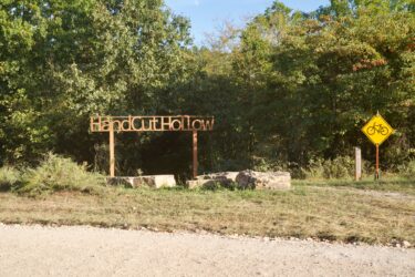 Sign marking the entrance to HandCut Hollow, surrounded by trees and shrubs, with a gravel path leading into the area. A yellow bike symbol sign is visible nearby. Hand Cut Hollow mountain bike trail.