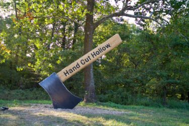 A large decorative axe sculpture with the text "Hand Cut Hollow" on the wooden handle, standing upright in a grassy area with trees in the background. Hand Cut Hollow mountain bike trail.