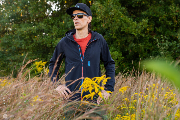 A person wearing a black jacket, red shirt, and sunglasses stands confidently in a field of tall grass and yellow flowers, with lush green trees in the background. The individual has long hair and a black cap, and appears focused and engaged with the natural environment.