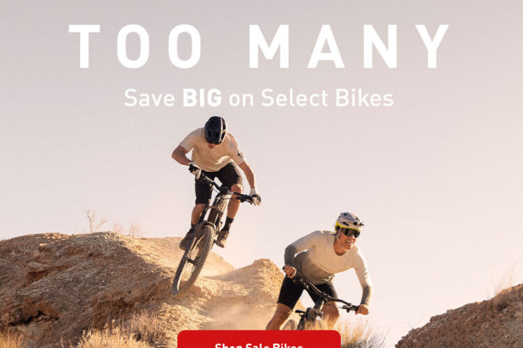 Two mountain bikers riding uphill on a rocky terrain, with dust clouds kicked up around them. The image features bold text overlay that reads "WE MADE TOO MANY" and "Save BIG on Select Bikes," along with a red button that says "Shop Sale Bikes." The background showcases a soft gradient sky.