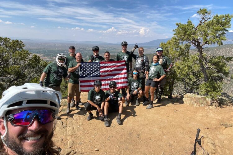 A group of ten individuals in mountain biking gear, standing on a rocky path with a scenic view in the background. They are smiling and posing for the camera, holding an American flag at the center. The group is wearing matching green shirts and includes both men and women. A mountain bike is visible on the ground nearby. The sky is clear with some clouds, and trees surround the area.
