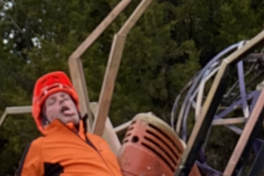 A person in an orange and black outfit and helmet is sitting on the ground, appearing exhausted or in a relaxed pose. They are surrounded by a few pieces of equipment, including a rusty red machine and wooden structures, with trees in the background under a cloudy sky. Lehigh Portland Trails mountain bike trail.